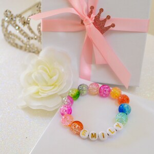 May include: A colorful beaded bracelet with the name "EMILY" spelled out in gold letters. The bracelet features a variety of iridescent beads in shades of pink, orange, green, and blue. A white gift box with a pink ribbon and a decorative crown is in the background.
