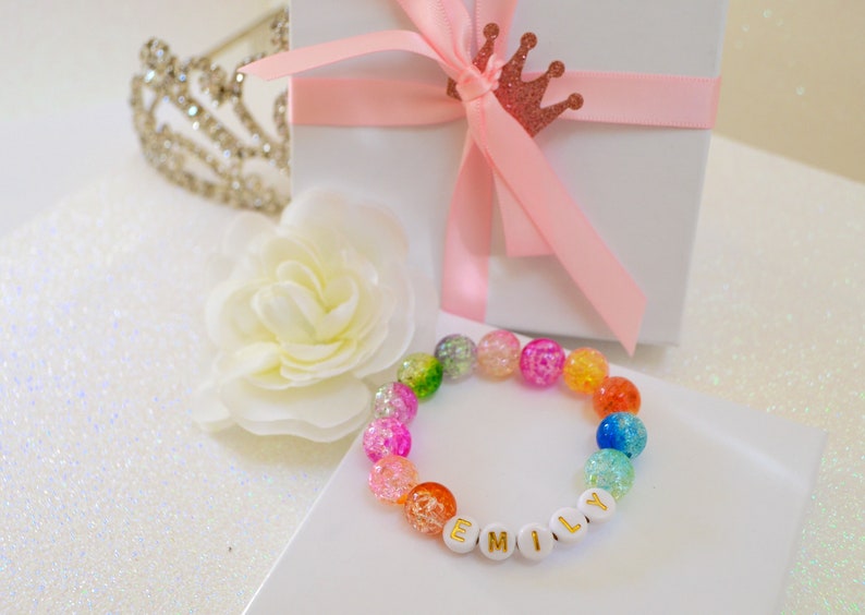 May include: A colorful beaded bracelet with the name "EMILY" spelled out in gold letters. The bracelet features a variety of iridescent beads in shades of pink, orange, green, and blue. A white gift box with a pink ribbon and a decorative crown is in the background.