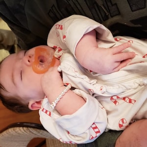 Puede incluir: Un beb&eacute; viste un pijama blanco de manga larga con un estampado de bastones de caramelo rojos y blancos. El beb&eacute; tiene un chupete naranja en la boca y lleva una pulsera de cuentas blancas con peque&ntilde;os detalles dorados en la mu&ntilde;eca izquierda.