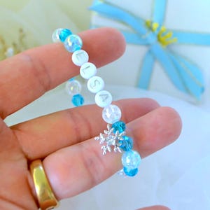 Könnte beinhalten: Ein blau-weißes Perlenarmband mit dem Namen "Elsa". Das Armband hat klare und blaue Perlen, einen silbernen Schneeflocken-Anhänger und wird in einer Hand gehalten. Ein goldener Ring ist ebenfalls sichtbar.