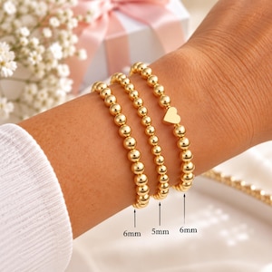 May include: Three gold-toned beaded bracelets worn together. Two bracelets feature 6mm beads, and one central bracelet features 5mm beads. The bracelet on the right includes a small, flat gold-toned heart charm. The beads are round and polished.