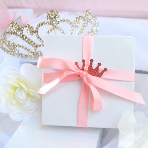 Puede incluir: Una caja de regalo cuadrada blanca atada con una cinta de sat&eacute;n rosa claro y adornada con un peque&ntilde;o dije de corona brillante en oro rosa. Una tiara de diamantes de imitaci&oacute;n met&aacute;licos se sit&uacute;a detr&aacute;s de la caja junto a una flor de tela.