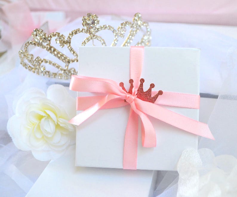 Puede incluir: Una caja de regalo cuadrada blanca atada con una cinta de sat&eacute;n rosa claro y adornada con un peque&ntilde;o dije de corona brillante en oro rosa. Una tiara de diamantes de imitaci&oacute;n met&aacute;licos se sit&uacute;a detr&aacute;s de la caja junto a una flor de tela.