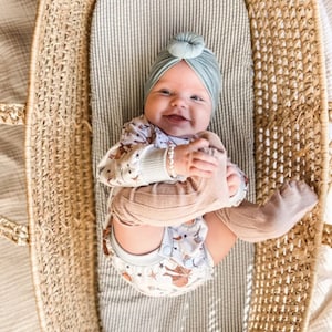Puede incluir: Un beb&eacute; con un turbante anudado de color verde salvia y un mameluco de manga larga con un estampado de polillas, soles y estrellas. El beb&eacute; tambi&eacute;n lleva calcetines acanalados de color tostado y una peque&ntilde;a pulsera de cuentas en un mois&eacute;s de mimbre.