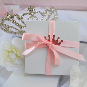 May include: A white gift box tied with a light pink ribbon and a glittery red crown embellishment. A silver tiara and a white flower are in the background, suggesting a theme of celebration or royalty.