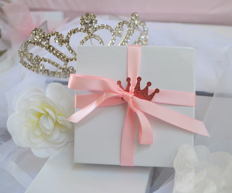 May include: A white gift box tied with a light pink ribbon and a glittery red crown embellishment. A silver tiara and a white flower are in the background, suggesting a theme of celebration or royalty.