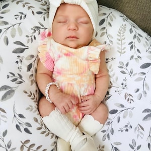 Puede incluir: Un beb&eacute; durmiendo con una diadema blanca acanalada con lazo, un mameluco acanalado tie-dye rosa y naranja con mangas con volantes, una pulsera de cuentas blanca con el nombre MOLLY, calcetines blancos con volantes y zapatos blancos.