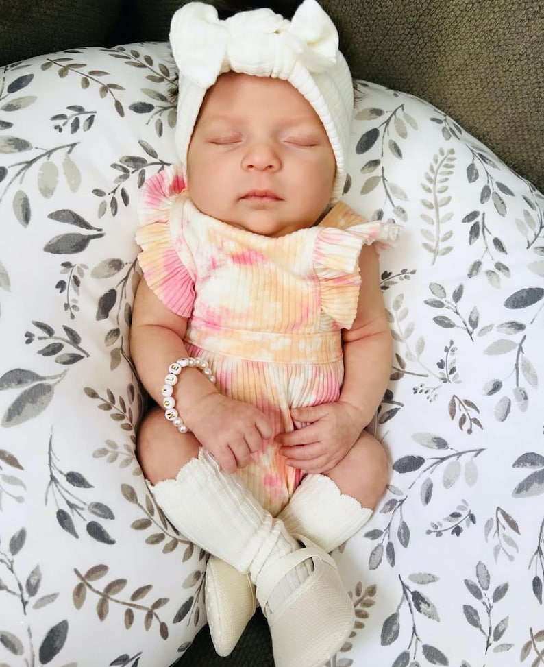 Puede incluir: Un beb&eacute; durmiendo con una diadema blanca acanalada con lazo, un mameluco acanalado tie-dye rosa y naranja con mangas con volantes, una pulsera de cuentas blanca con el nombre MOLLY, calcetines blancos con volantes y zapatos blancos.