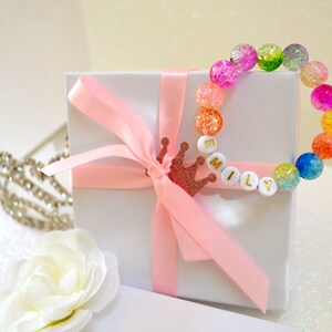 May include: A colorful beaded bracelet with the name "MILY" spelled out in white letter beads. The bracelet is adorned with a variety of crackle beads in shades of pink, green, orange, and blue. A white gift box is tied with a pink ribbon and a glittery crown.