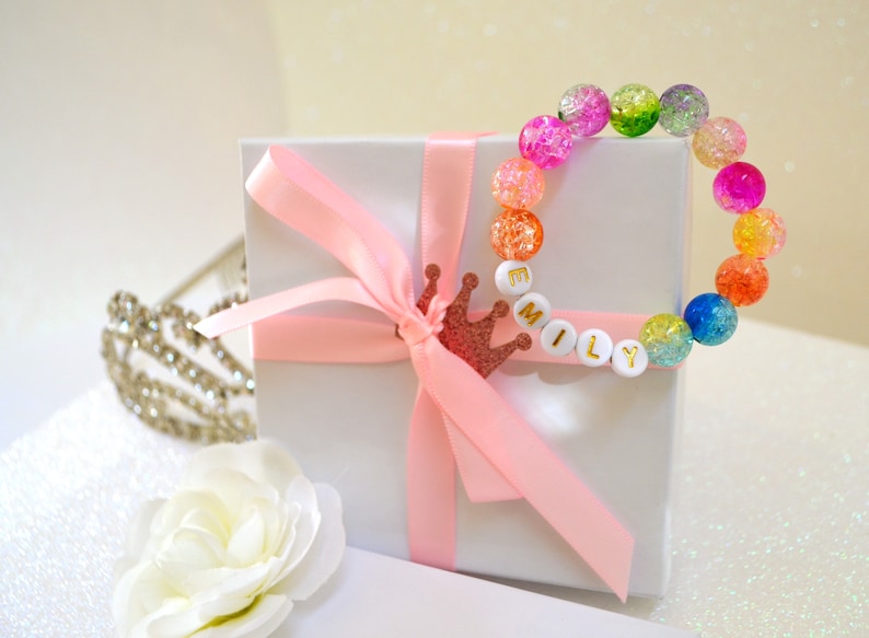 May include: A colorful beaded bracelet with the name "MILY" spelled out in white letter beads. The bracelet is adorned with a variety of crackle beads in shades of pink, green, orange, and blue. A white gift box is tied with a pink ribbon and a glittery crown.