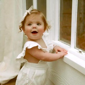 Puede incluir: Un ni&ntilde;o peque&ntilde;o con un pelele blanco sin mangas con tirantes con volantes y un lazo para el pelo blanco a juego. El pelele presenta una cintura ajustada y un dise&ntilde;o sencillo. Se puede ver una peque&ntilde;a pulsera de cuentas en la mu&ntilde;eca.