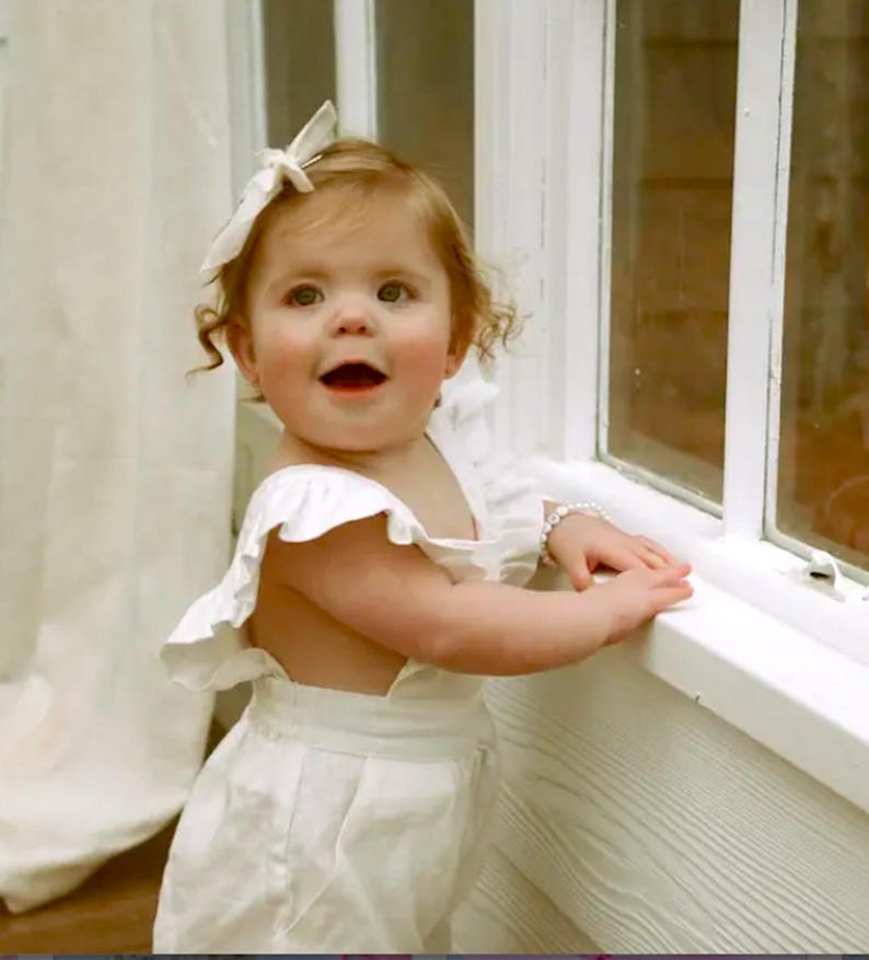 Puede incluir: Un ni&ntilde;o peque&ntilde;o con un pelele blanco sin mangas con tirantes con volantes y un lazo para el pelo blanco a juego. El pelele presenta una cintura ajustada y un dise&ntilde;o sencillo. Se puede ver una peque&ntilde;a pulsera de cuentas en la mu&ntilde;eca.