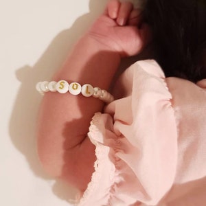 Puede incluir: Una pulsera de cuentas con esferas blancas y cuentas redondas blancas con letras doradas que forman la palabra SOL. La pulsera se lleva en el brazo, destacando las cuentas con letras entre las cuentas blancas y de aspecto perlado.