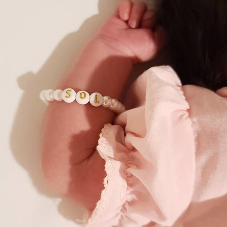 Puede incluir: Una pulsera de cuentas con esferas blancas y cuentas redondas blancas con letras doradas que forman la palabra SOL. La pulsera se lleva en el brazo, destacando las cuentas con letras entre las cuentas blancas y de aspecto perlado.