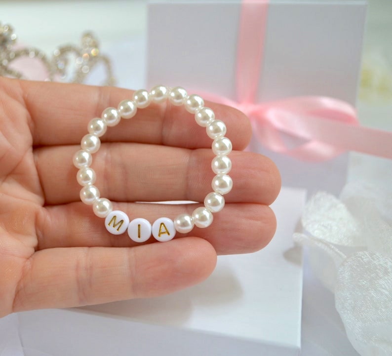 Puede incluir: Una pulsera circular compuesta por cuentas de perlas blancas y tres cuentas esf&eacute;ricas blancas con las letras doradas M, I y A. La pulsera se sostiene en una palma, con una caja de regalo blanca y una cinta rosa visibles al fondo.