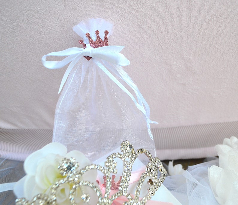 May include: A white organza gift bag with a rose gold glitter crown and white ribbon. A silver tiara with clear rhinestones and white flowers are in the foreground. The background is a light pink fabric.