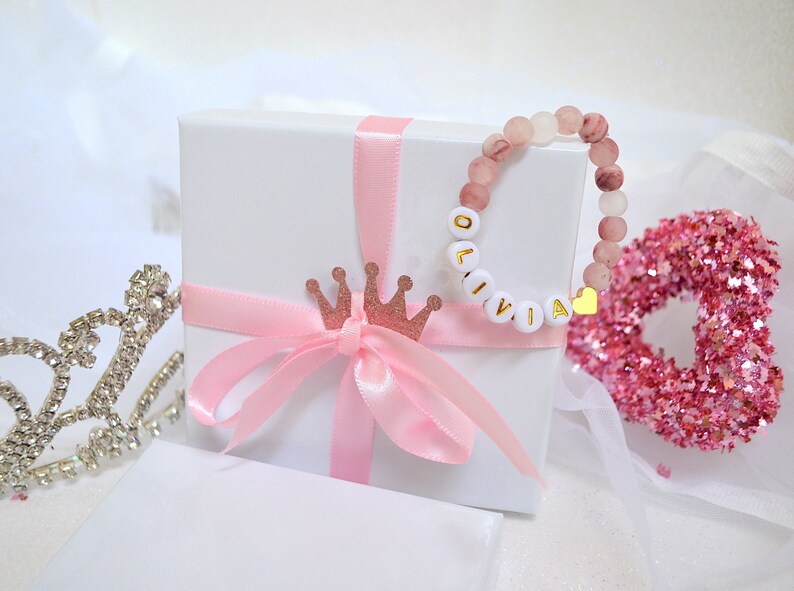 Pode incluir: Uma caixa de presente branca com uma fita rosa e uma coroa dourada. A caixa est&aacute; ao lado de uma pulseira de contas rosa e branca com o nome "Olivia" escrito nela.