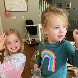 May include: Two young children are smiling. One child is wearing a pink and white dress and waving. The other child is wearing a teal long-sleeve shirt with a rainbow graphic. A green toy is in the child's hand.
