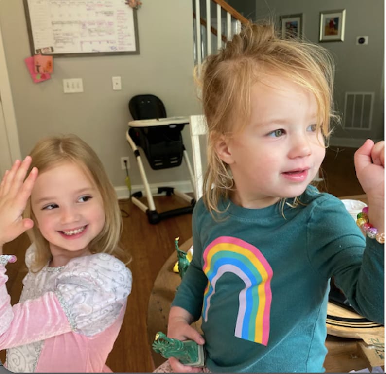 May include: Two young children are smiling. One child is wearing a pink and white dress and waving. The other child is wearing a teal long-sleeve shirt with a rainbow graphic. A green toy is in the child's hand.