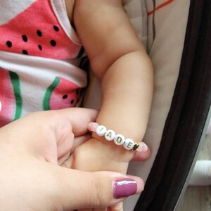 Pode incluir: Um bracelete de contas brancas com letras douradas que soletram o nome "JADE". O bracelete tem um pequeno fecho de cora&ccedil;&atilde;o dourado.