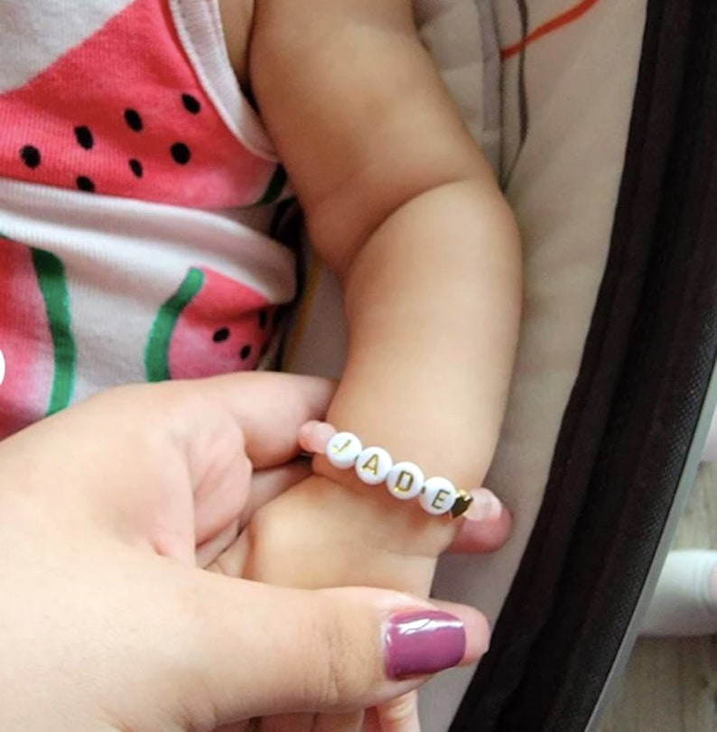 Pode incluir: Um bracelete de contas brancas com letras douradas que soletram o nome "JADE". O bracelete tem um pequeno fecho de cora&ccedil;&atilde;o dourado.