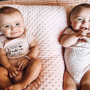 Pode incluir: Dois beb&ecirc;s vestindo macac&otilde;es brancos com bolinhas pretas est&atilde;o deitados em um travesseiro estampado em rosa e branco. Um macac&atilde;o tem o texto "You make me smile!" bordado.