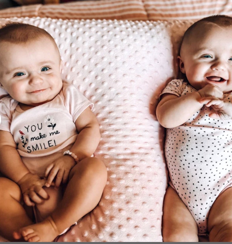 Pode incluir: Dois beb&ecirc;s vestindo macac&otilde;es brancos com bolinhas pretas est&atilde;o deitados em um travesseiro estampado em rosa e branco. Um macac&atilde;o tem o texto "You make me smile!" bordado.
