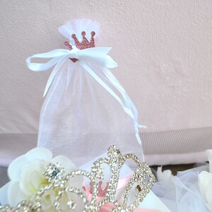 Puede incluir: Una bolsa de regalo de organza blanca transparente con cord&oacute;n, que presenta un peque&ntilde;o recorte de corona con purpurina en oro rosa en la parte delantera. La bolsa est&aacute; atada con una cinta de sat&eacute;n blanca. En primer plano, una tiara de metal plateado.