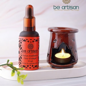 Peut inclure: Une bouteille en verre marron avec un compte-gouttes et une étiquette rouge avec le texte "be artisan" et "Parfum d'ambiance Fleur d'oranger 50 ml". À côté, un brûleur d'huile en céramique marron avec une bougie allumée. Sur un plateau blanc.