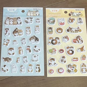 May include: Two sticker sheets featuring various cat illustrations in different poses and colors. The stickers are on light blue and yellow backgrounds, with Japanese text at the top. The stickers include cats with donuts, ice cream, and other treats.