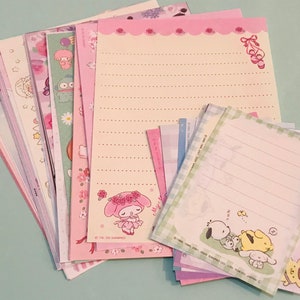 May include: A stack of pink and green stationery paper with various cartoon characters and designs. The paper is lined and has a decorative border.