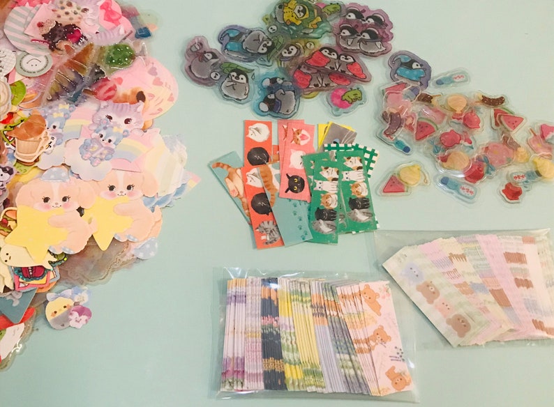 May include: Assortment of colorful stickers featuring various cartoon animals, including cats, penguins, and bears. The stickers are arranged in three groups on a light blue surface.