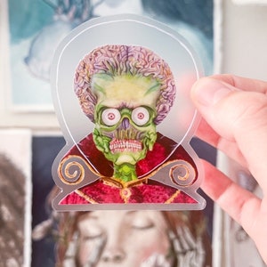 May include: A clear sticker with a cartoon image of a green alien with a large brain, wearing a red and gold robe. The alien has large eyes and a toothy grin.