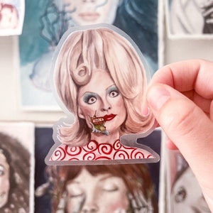Puede incluir: Una pegatina transparente con un retrato estilizado de una mujer con cabello rubio y lápiz labial rojo. El rostro de la mujer tiene una apariencia surrealista, parcialmente descompuesta. La pegatina se sostiene en una mano, con otras obras de arte visibles en el fondo.