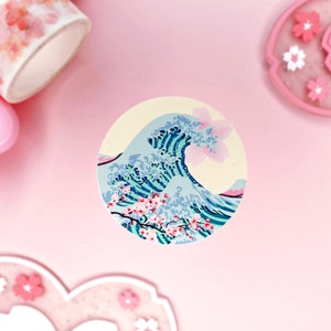 May include: A round sticker featuring a blue and green wave design, inspired by Japanese art, with pink cherry blossoms on a cream background.