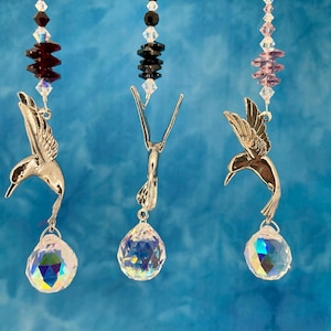 May include: Three hummingbird suncatchers with iridescent crystal teardrop pendants. Each suncatcher features a silver-colored hummingbird charm and is strung with colorful beads. The background is a gradient of blue.