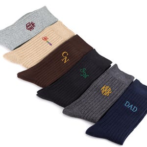 K&ouml;nnte beinhalten: Ein Set aus sechs gerippten Socken in verschiedenen Farben, darunter Hellgrau, Beige, Braun, Schwarz, Anthrazit und Marineblau. Jede Socke ist mit gesticktem Text oder Initialen versehen, namentlich "OUR", "Groom", "GN", "SM", ein Monogramm und "DAD".