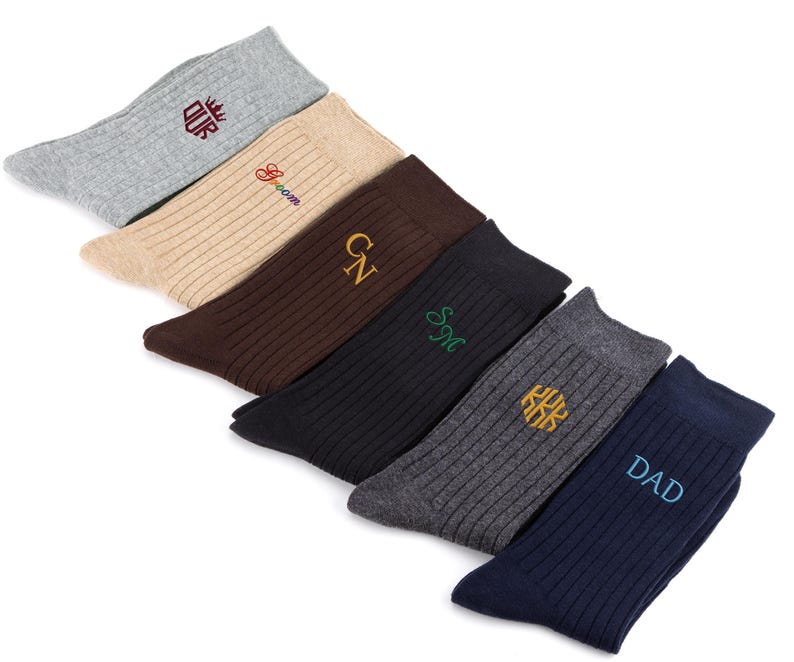 K&ouml;nnte beinhalten: Ein Set aus sechs gerippten Socken in verschiedenen Farben, darunter Hellgrau, Beige, Braun, Schwarz, Anthrazit und Marineblau. Jede Socke ist mit gesticktem Text oder Initialen versehen, namentlich "OUR", "Groom", "GN", "SM", ein Monogramm und "DAD".