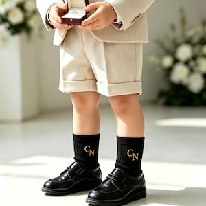 May include: A beige blazer and matching shorts paired with black ribbed socks featuring gold embroidered CN initials and black leather dress shoes. A small burgundy box containing a ring is held in the center of the frame.