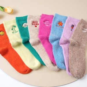 May include: A collection of colorful socks, each with unique embroidered designs and text. The socks are in shades of orange, yellow, teal, beige, pink, blue, and lavender. Some socks feature a smiley face, a palm tree, a dog, a flower, and text that reads "BEST MAMA" and "OUR NAME".