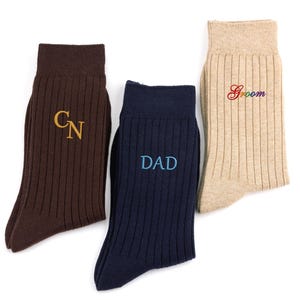 K&ouml;nnte beinhalten: Drei Paar gerippte Socken in Braun, Marineblau und Beige. Die braune Socke zeigt die gold gestickten Buchstaben CN, die marineblaue Socke den hellblau gestickten Text DAD und die beige Socke das Wort Groom in Regenbogenfarben.