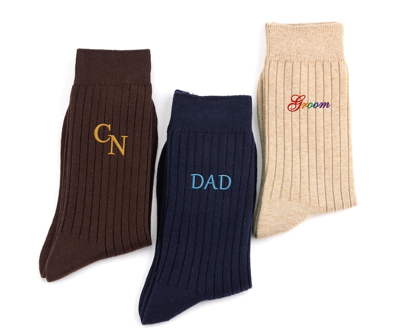 K&ouml;nnte beinhalten: Drei Paar gerippte Socken in Braun, Marineblau und Beige. Die braune Socke zeigt die gold gestickten Buchstaben CN, die marineblaue Socke den hellblau gestickten Text DAD und die beige Socke das Wort Groom in Regenbogenfarben.