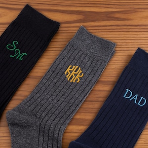 K&ouml;nnte beinhalten: Drei Paar gerippte Socken in Schwarz, Anthrazit und Marineblau. Die schwarze Socke zeigt gr&uuml;ne gestickte Initialen SM, die anthrazitfarbene Socke ein goldenes gesticktes hexagonales Monogramm KKK und die marineblaue Socke den blauen gestickten Text DAD.