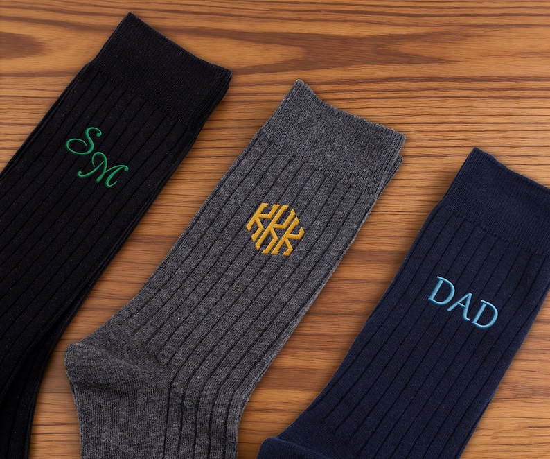 K&ouml;nnte beinhalten: Drei Paar gerippte Socken in Schwarz, Anthrazit und Marineblau. Die schwarze Socke zeigt gr&uuml;ne gestickte Initialen SM, die anthrazitfarbene Socke ein goldenes gesticktes hexagonales Monogramm KKK und die marineblaue Socke den blauen gestickten Text DAD.