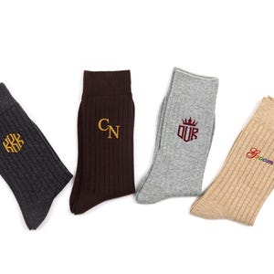 K&ouml;nnte beinhalten: Vier Paar gerippte Socken in Anthrazit, Dunkelbraun, Hellgrau und Beige. Die anthrazitfarbene Socke hat ein goldenes sechseckiges Monogramm, die braune Socke goldene Initialen CN, die graue Socke ein kastanienbraunes Kronenlogo mit OUR und die beige Socke das Wort Groom in Regenbogenfarben.