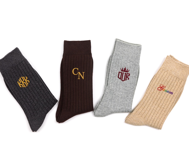 K&ouml;nnte beinhalten: Vier Paar gerippte Socken in Anthrazit, Dunkelbraun, Hellgrau und Beige. Die anthrazitfarbene Socke hat ein goldenes sechseckiges Monogramm, die braune Socke goldene Initialen CN, die graue Socke ein kastanienbraunes Kronenlogo mit OUR und die beige Socke das Wort Groom in Regenbogenfarben.
