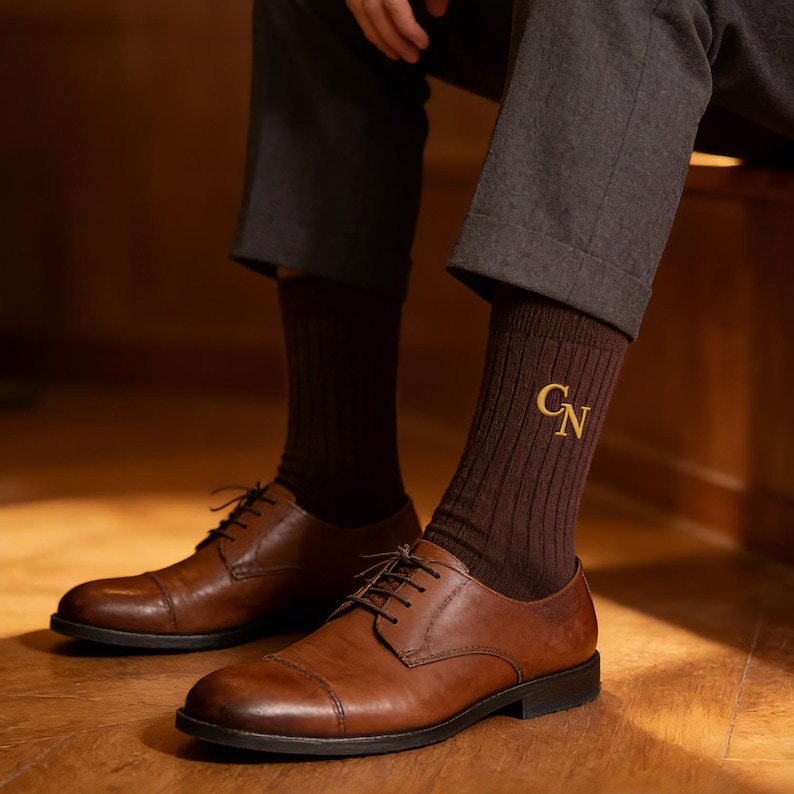 K&ouml;nnte beinhalten: Ein Paar dunkelbraune Rippsocken mit den gestickten goldenen Buchstaben CN an der Seite, kombiniert mit dunkelbraunen Lederschuhen mit Kappennaht und einer dunkelgrauen Hose. Die Socken haben eine vertikale Rippstruktur und mittlere L&auml;nge.