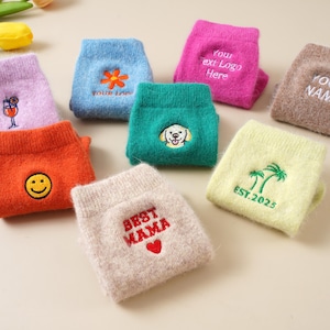 Può includere: Una collezione di calzini colorati ricamati. I calzini sono in vari colori, tra cui arancione, blu, rosa, verde e beige. Ogni calzino presenta un design unico, come una faccina sorridente, un fiore, un cane o testo come "Best Mama."