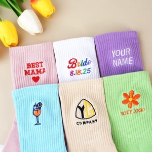 May include: Assorted colorful socks with embroidered designs and text. The socks include the phrases "BEST MAMA", "Bride 8.18.25", and "YOUR NAME". Other socks feature a cocktail, a flower, and a company logo.