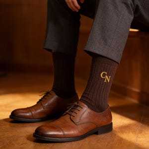 May include: Brown ribbed socks with gold embroidered initials "CN" are paired with brown leather dress shoes. The shoes have a classic cap-toe design and are laced up. The person is wearing gray trousers.